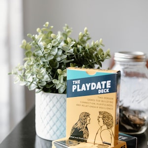 Op de afbeelding: The Playdate Deck, een spel voor twee personen om verbinding, speelsheid en relatie-veerkracht op te bouwen. De doos is geel met een turquoise bovenkant en bevat illustraties van twee mensen. Een witte pot met patroon en een groene plant staat op de achtergrond.