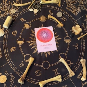May include: A black and gold zodiac-themed altar cloth with bones, coins, and a card that reads "Surrender". The card has a red and pink design. Other items include a cork and a small green figurine.