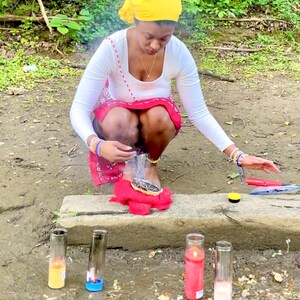 Può includere: Una persona con un foulard giallo e una camicia bianca a maniche lunghe è inginocchiata vicino a uno specchio d'acqua. Diverse candele accese in contenitori di vetro sono poste vicino all'acqua. Sono presenti anche un panno rosso e un coltello.