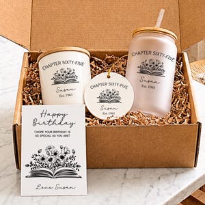 May include: A gift box containing a white candle with a wooden lid, a round ceramic ornament, and a frosted glass tumbler with a bamboo lid and straw. Each item features the text "CHAPTER SIXTY-FIVE," "Susan," "Est. 1961," and a floral book graphic.
