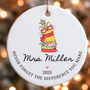 Puede incluir: Adorno navide&ntilde;o redondo de cer&aacute;mica blanca con un gato de dibujos animados con gafas de sol en forma de coraz&oacute;n y una bufanda. El adorno tiene el texto "Mrs. Miller", "Never Forget the Difference You Make" y el a&ntilde;o 2025.