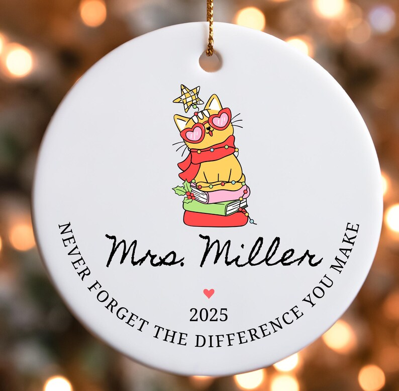 Puede incluir: Adorno navide&ntilde;o redondo de cer&aacute;mica blanca con un gato de dibujos animados con gafas de sol en forma de coraz&oacute;n y una bufanda. El adorno tiene el texto "Mrs. Miller", "Never Forget the Difference You Make" y el a&ntilde;o 2025.