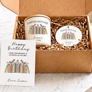 May include: A gift box containing a white candle and a round ceramic ornament, both featuring a graphic of books with flowers and the text 'CHAPTER SIXTY-FIVE Susan Est. 1961'. A greeting card reads 'Happy Birthday' with the same book graphic.