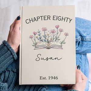 May include: A beige book cover with the text "CHAPTER EIGHTY" above a floral illustration and the name "Susan" in cursive. Below is "Est. 1946". The book is held by a person wearing a blue sweater and jeans.