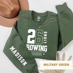 Puede incluir: Sudadera verde militar con texto blanco. La parte delantera muestra "26 ROWING LIONS SENIOR" y la manga "MADISON". También se ven un collar dorado y un sombrero marrón.