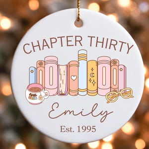 May include: A white ceramic ornament with a gold string for hanging. The ornament features the text "CHAPTER THIRTY" above a row of illustrated books, a teacup, and glasses. The name "Emily" and "Est. 1995" are also displayed.