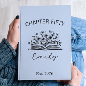 May include: A light blue journal with the text "CHAPTER FIFTY" above a floral illustration and the name "Emily" below. The journal also includes the text "Est. 1976". The journal is held by a person.