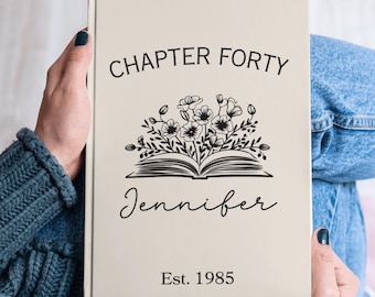 May include: A beige journal with the text "CHAPTER FORTY" above a floral illustration and the name "Jennifer" below. The journal also includes the text "Est. 1985".