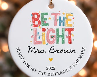 May include: A white ceramic ornament with the text "BE THE LIGHT" in colorful letters, decorated with string lights and stars. The name "Mrs. Brown" is written below, along with "2025" and a gold heart. The phrase "NEVER FORGET THE DIFFERENCE YOU MAKE" is written around the edge.