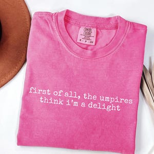 May include: A hot pink t-shirt with the text "first of all, the umpires think i'm a delight" in white. The shirt has a crew neck and a Comfort Colors tag. The shirt is folded and displayed on a white surface.