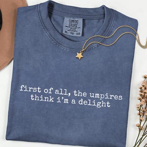 May include: A blue Comfort Colors t-shirt with the text "first of all, the umpires think i'm a delight" in white. A gold star pendant necklace rests on the shirt. The shirt is size XL.