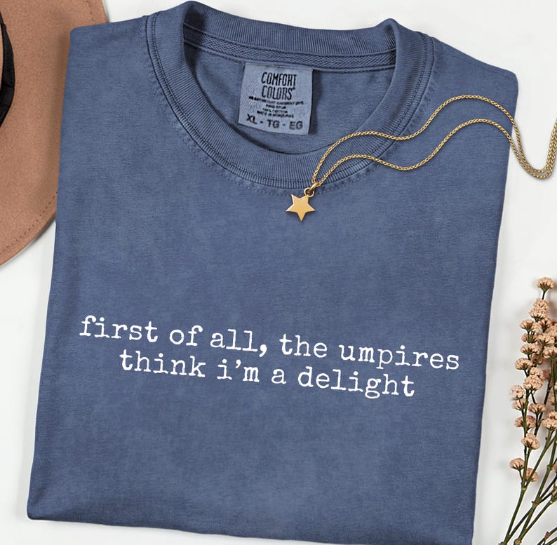May include: A blue Comfort Colors t-shirt with the text "first of all, the umpires think i'm a delight" in white. A gold star pendant necklace rests on the shirt. The shirt is size XL.