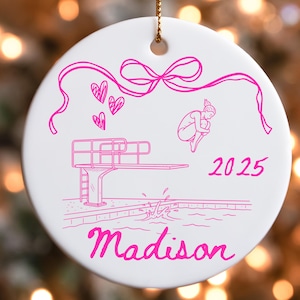 May include: White ceramic ornament with a pink illustration of a diver, diving board, hearts, and a bow. The text "2025" and "Madison" are also in pink. A gold string is attached for hanging.