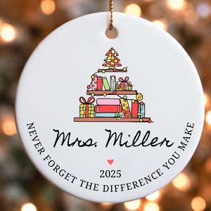 Puede incluir: Adorno navide&ntilde;o redondo de cer&aacute;mica blanca con una cuerda dorada. El adorno presenta una colorida ilustraci&oacute;n de un &aacute;rbol de Navidad hecho de libros y regalos. El texto incluye "Mrs. Miller", un coraz&oacute;n y "2025".
