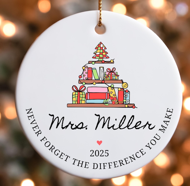 Puede incluir: Adorno navide&ntilde;o redondo de cer&aacute;mica blanca con una cuerda dorada. El adorno presenta una colorida ilustraci&oacute;n de un &aacute;rbol de Navidad hecho de libros y regalos. El texto incluye "Mrs. Miller", un coraz&oacute;n y "2025".