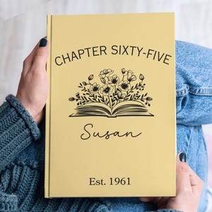 May include: A yellow journal with the text "CHAPTER SIXTY-FIVE" and the name "Susan" in a script font. A floral design is above the name. The journal also has the text "Est. 1961".