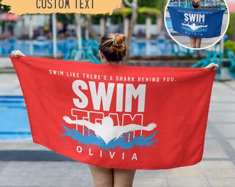 May include: A red swim team towel with the text "Swim Like There's a Shark Behind You." The towel features a swimmer graphic and the words "Swim Team" and "Olivia" in white and blue. The towel is held up by a person.
