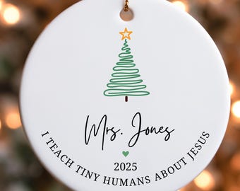 May include: White ceramic Christmas ornament with a green and gold Christmas tree design. The ornament has the text "Mrs. Jones" and "I teach tiny humans about Jesus 2025". A gold string is attached for hanging.
