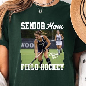 May include: A dark green t-shirt featuring a graphic print with the text SENIOR Mom and FIELD HOCKEY. The graphic includes a photograph of a field hockey player in a navy uniform with the number 7, and the name Olivia 2027 in white script.