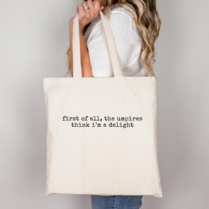 May include: Cream-colored tote bag with the text "first of all, the umpires think i'm a delight" printed in black. The bag has two handles and is held by a person. The bag is made of a canvas-like material.
