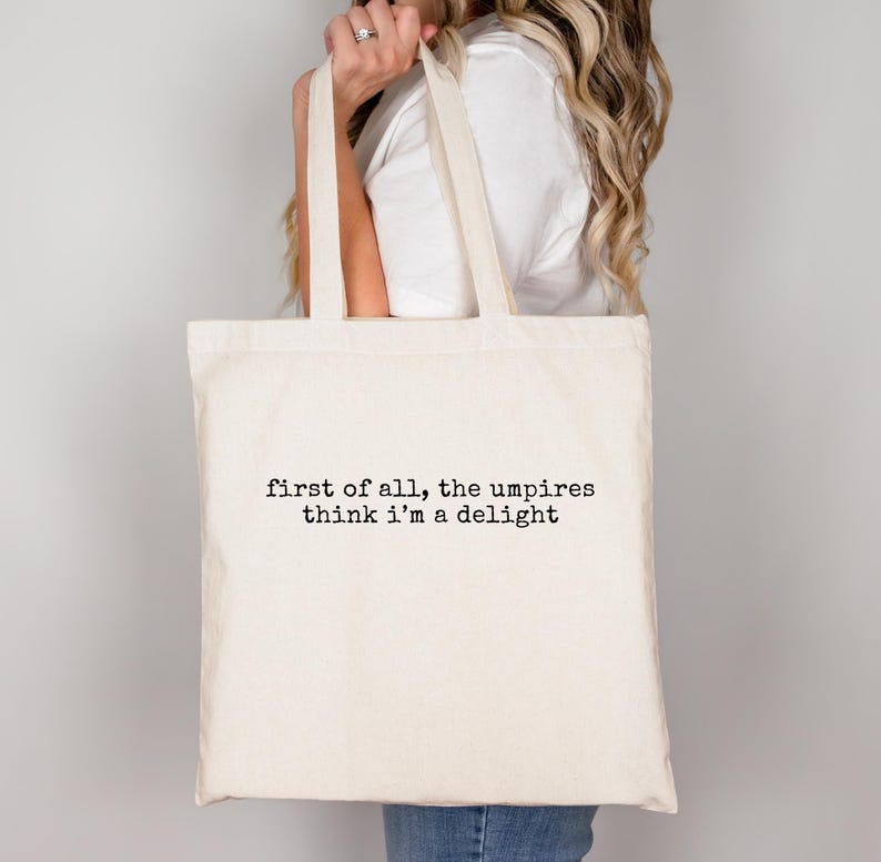 May include: Cream-colored tote bag with the text "first of all, the umpires think i'm a delight" printed in black. The bag has two handles and is held by a person. The bag is made of a canvas-like material.