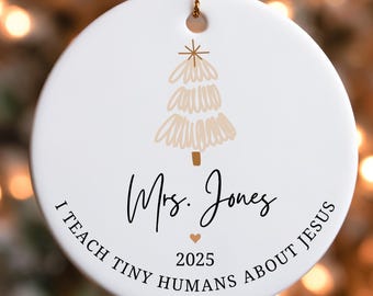 May include: A white ceramic Christmas ornament with a gold string. The ornament features a beige Christmas tree design and the text "Mrs. Jones", "I TEACH TINY HUMANS ABOUT JESUS", and the year "2025".