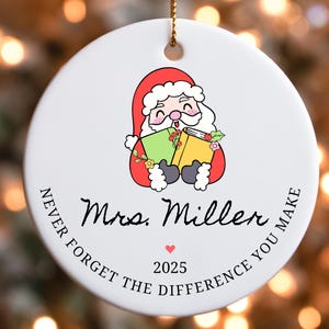 Puede incluir: Adorno navide&ntilde;o redondo de cer&aacute;mica blanca con una ilustraci&oacute;n de Pap&aacute; Noel sosteniendo libros. El adorno incluye el texto "Mrs. Miller", un coraz&oacute;n, el a&ntilde;o "2025" y la frase "Never forget the difference you make".
