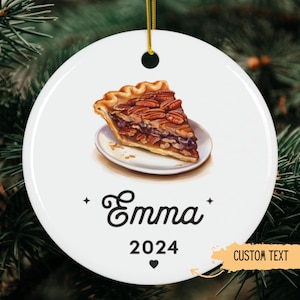 May include: White ceramic ornament with a slice of pecan pie on a small plate. The ornament has the name "Emma" and the year "2024" printed on it, with a heart symbol. The words "CUSTOM TEXT" are also visible.