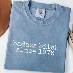 May include: A blue Comfort Colors t-shirt with the text "badass bitch since 1976" in white. The shirt is a light blue color and has a vintage, worn look. The shirt is folded neatly on a white surface.