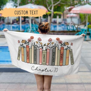 May include: A white beach towel with a row of colorful books with floral accents. The text "Chapter 70" is printed below the books. The words "Timeless", "Linda", "Historical", "Iconic", "Vintage", "1955", and "Masterpiece" are printed on the books.