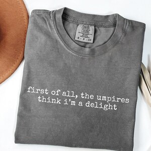 May include: A gray Comfort Colors t-shirt with the text "first of all, the umpires think i'm a delight" in white. The shirt is folded and has a small tag at the neckline. A brown hat is partially visible in the upper left corner.