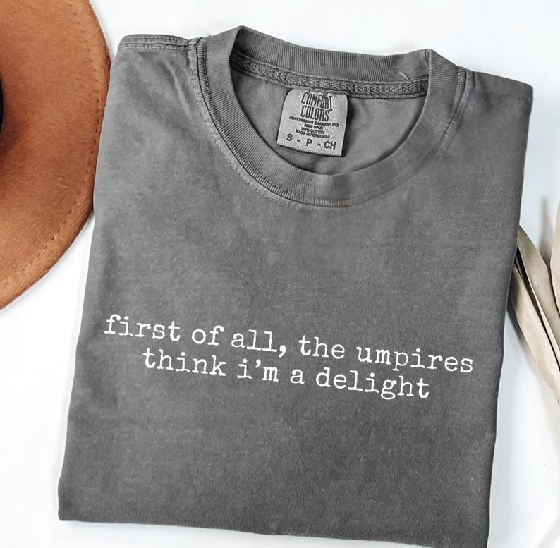 May include: A gray Comfort Colors t-shirt with the text "first of all, the umpires think i'm a delight" in white. The shirt is folded and has a small tag at the neckline. A brown hat is partially visible in the upper left corner.
