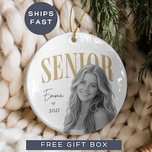 May include: A round white ceramic ornament featuring the gold text SENIOR, the name Emma in black script, a small gold heart, and the year 2027. A black and white portrait of a young person with long hair is printed on the front. It hangs by a gold cord.