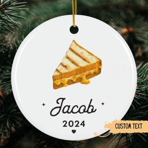 May include: White ceramic ornament with a cartoon illustration of a grilled cheese sandwich. The ornament has the name "Jacob" and the year "2024" printed on it. The text "CUSTOM TEXT" is also visible on the ornament.