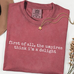 May include: A dusty rose Comfort Colors t-shirt with the text "first of all, the umpires think i'm a delight" in white. A gold star pendant necklace rests on the shirt. The shirt is size XL-TG-EG.