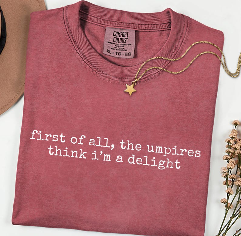 May include: A dusty rose Comfort Colors t-shirt with the text "first of all, the umpires think i'm a delight" in white. A gold star pendant necklace rests on the shirt. The shirt is size XL-TG-EG.