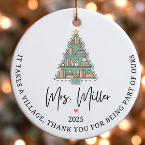 Puede incluir: Adorno navide&ntilde;o redondo de cer&aacute;mica blanca con una cuerda dorada. El adorno presenta un dise&ntilde;o de &aacute;rbol de Navidad hecho de libros, un mu&ntilde;eco de nieve y una estrella. El texto incluye "Mrs. Miller", "2025" y "It takes a village, thank you for being part of ours."