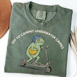 May include: Olive green Comfort Colors t-shirt featuring a cartoon frog riding a scooter, wearing a blue helmet and holding a yellow ball. The text "OFF TO COMMIT UNDERWATER CRIMES" is arched above the frog. The shirt has a crew neck and short sleeves.