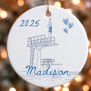 May include: White ceramic ornament with a blue illustration of a diver on a diving board, the year 2025, and the name "Madison." The diver is wearing a blue swimsuit. Three blue hearts are also drawn on the ornament.