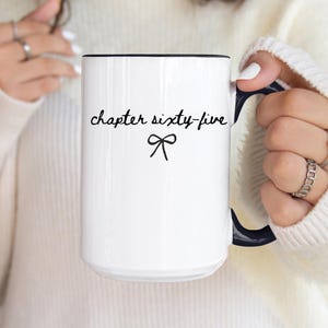 May include: White ceramic mug with a black rim and handle. The mug features the text "chapter sixty-five" in a handwritten font, with a small bow graphic below. The mug is held by a person wearing a white sweater.