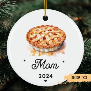 May include: White ceramic ornament with a watercolor illustration of an apple pie. The ornament has the word "Mom" in cursive, the year "2024", and a heart. A gold string is attached for hanging.