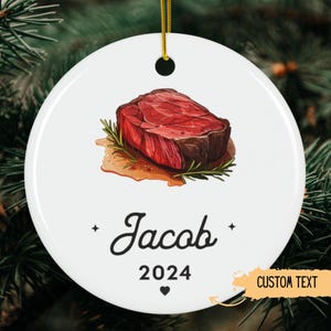 May include: White ceramic ornament with a cartoon illustration of a red steak on a bed of rosemary. The ornament has the text "Jacob 2024" and "CUSTOM TEXT" printed on it.