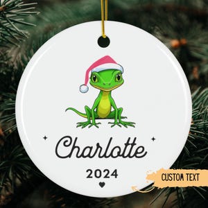 Puede incluir: Un adorno navideño de cerámica blanca con un lagarto verde que lleva un gorro de Papá Noel rosa. El adorno tiene el nombre "Charlotte" y el año "2024" impreso en él. El adorno está decorado con un pequeño corazón.