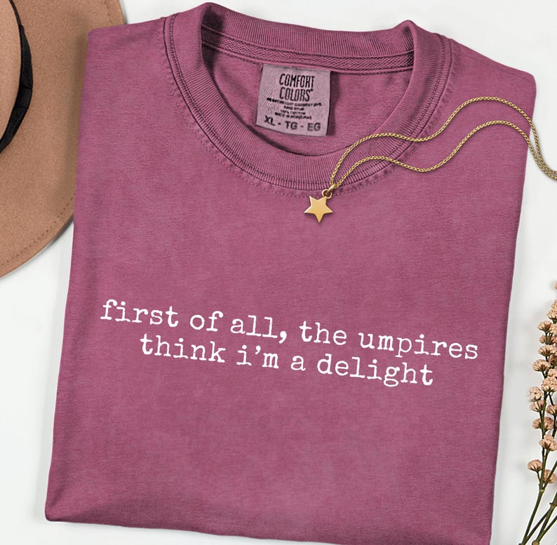 May include: A mauve-colored t-shirt with the text "first of all, the umpires think i'm a delight" in white. A gold star pendant necklace rests on the shirt. The shirt has a Comfort Colors tag, size XL.