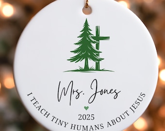 May include: White ceramic Christmas ornament with a green pine tree and cross design. The ornament has the text "Mrs. Jones", "I TEACH TINY HUMANS ABOUT JESUS", and the year "2025". A gold string is attached for hanging.