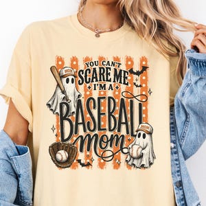 May include: A pale yellow t-shirt featuring a graphic with orange vertical stripes and white polka dots. The text reads: YOU CAN'T SCARE ME I'M A BASEBALL MOM. The design includes two ghosts wearing baseball caps, a baseball bat, a glove, and a ball.