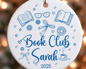 May include: White ceramic ornament with a gold string for hanging. The ornament features blue illustrations of books, glasses, bows, flowers, hearts, stars, and a coffee cup. The text reads "Book Club Sarah 2025."
