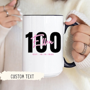 May include: White ceramic mug with a black interior and handle. The mug features the number "100" in black, with the name "Ellen" in pink script, and the date "EST. 10.10.1925". The words "CUSTOM TEXT" are at the bottom.