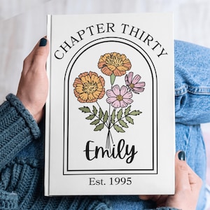 May include: A white journal with the text "CHAPTER THIRTY" arched above a floral illustration. The illustration features orange and pink flowers with green leaves. The name "Emily" is written below the flowers, with "Est. 1995" at the bottom.