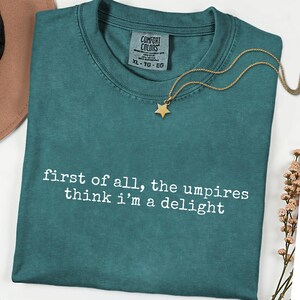 May include: Teal Comfort Colors t-shirt with the text "first of all, the umpires think i'm a delight" in white. A gold star pendant necklace rests on the shirt. The shirt has a crew neck and a tag that says XL-TG-EG.